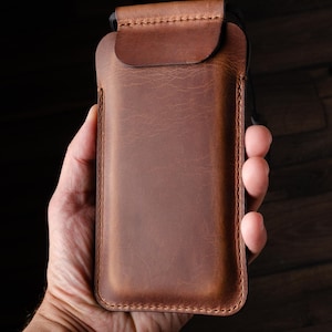 May include: A brown leather phone case with a flap closure. The case is held in a hand.