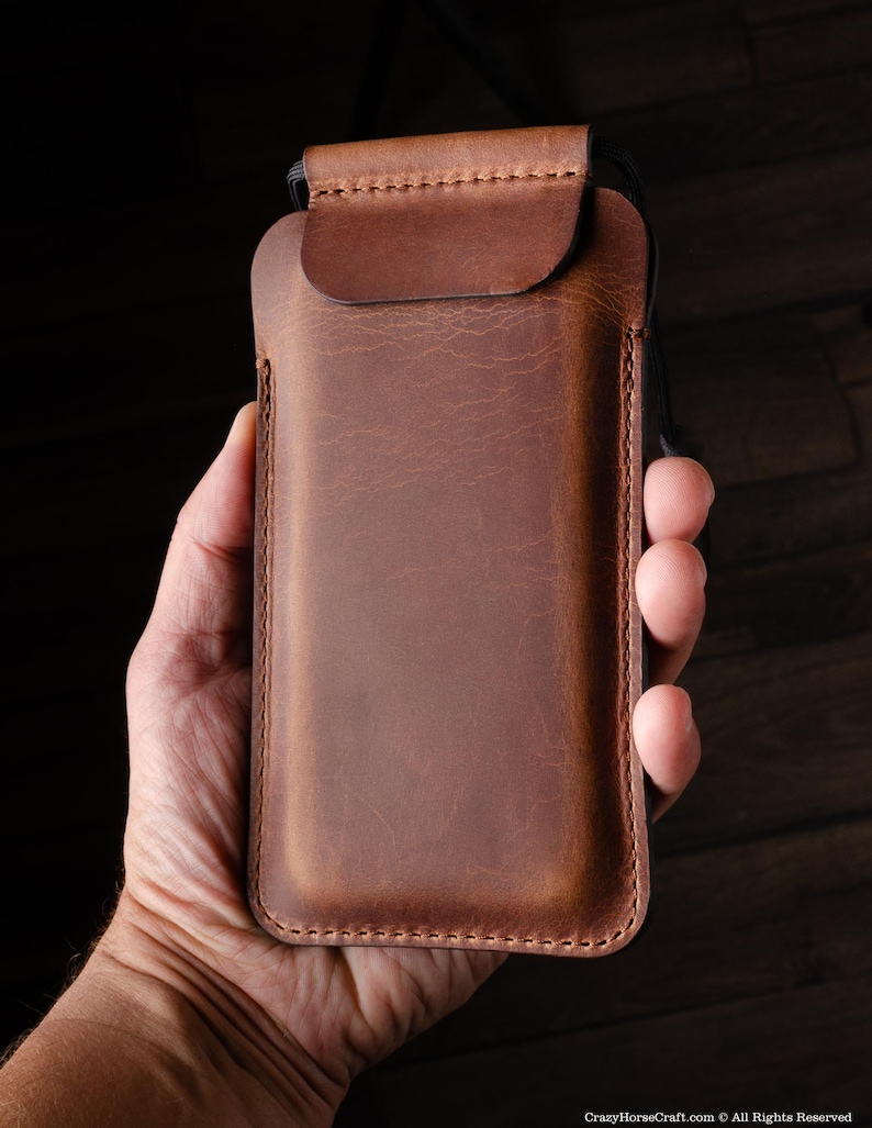 May include: A brown leather phone case with a flap closure. The case is held in a hand.