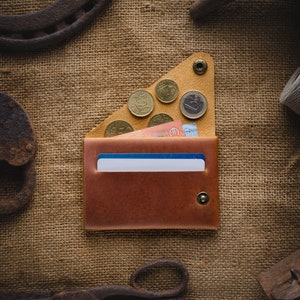 May include: A brown leather wallet with a flap closure and a snap button. The wallet is open and has a few euro coins and bills inside. The wallet is sitting on a burlap background.