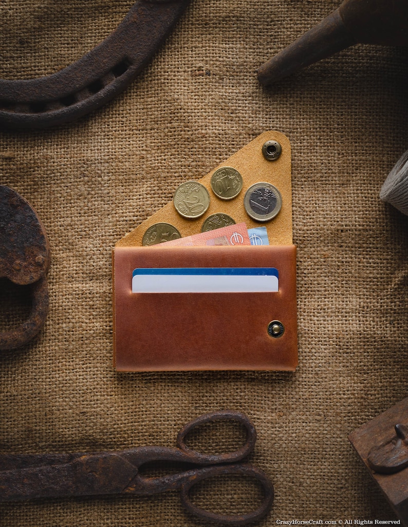 May include: A brown leather wallet with a flap closure and a snap button. The wallet is open and has a few euro coins and bills inside. The wallet is sitting on a burlap background.