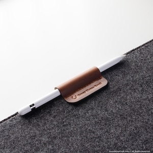 May include: A brown leather pencil holder with the text "Crazy Horse Craft" attached to a gray felt surface. A white Apple Pencil is held in the holder.