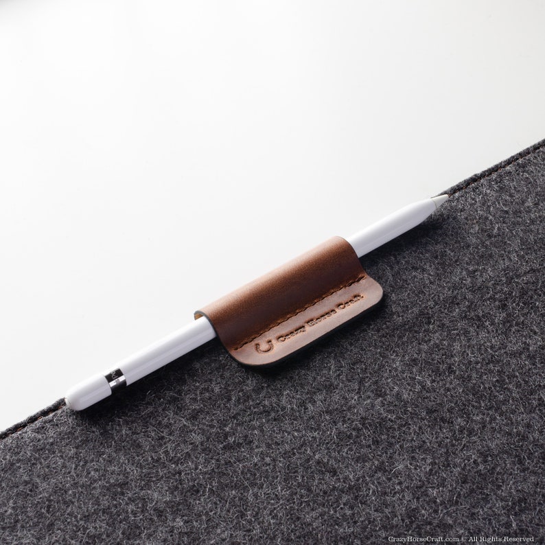 May include: A brown leather pencil holder with the text "Crazy Horse Craft" attached to a gray felt surface. A white Apple Pencil is held in the holder.