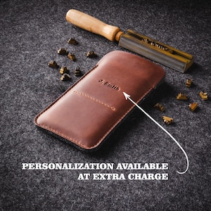 May include: A brown leather phone case with the name "J. Smith" embossed on the front. The case is lying on a gray surface with a metal tool and brass letters scattered around it. The text "PERSONALIZATION AVAILABLE AT EXTRA CHARGE" is printed below the case.
