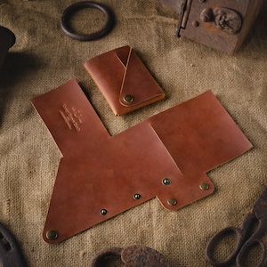 May include: A brown leather wallet with a fold-over flap and a snap closure. The wallet has a small pocket on the front and is decorated with brass rivets. The wallet is lying on a burlap surface.