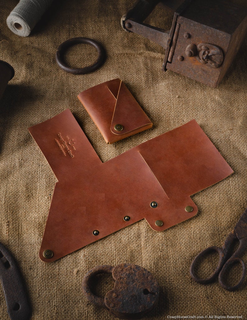 May include: A brown leather wallet with a fold-over flap and a snap closure. The wallet has a small pocket on the front and is decorated with brass rivets. The wallet is lying on a burlap surface.