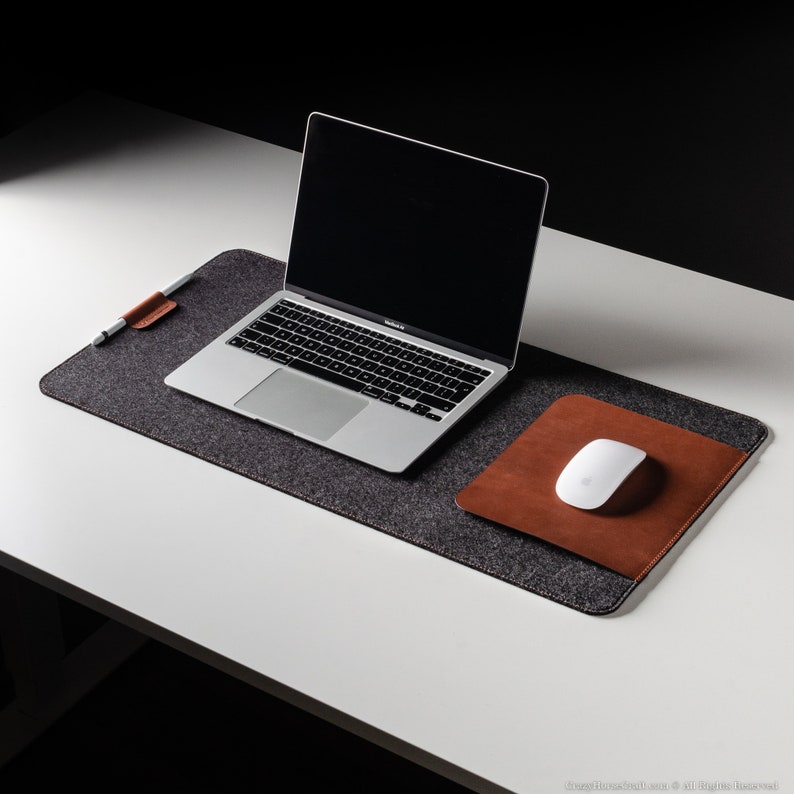 May include: A gray felt desk mat with a brown leather mouse pad and a silver laptop computer on top. The laptop is open and the screen is black. A white wireless mouse is on the brown leather mouse pad.