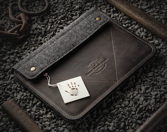 May include: A brown leather laptop sleeve with a gray felt flap and a small tag with a handprint logo. The sleeve is on a bed of small gray rocks.