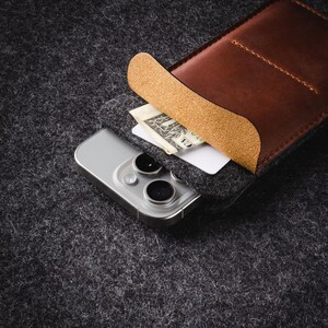May include: A brown leather phone case with a flap that folds over the top of the phone. The case is lined with a tan fabric and has a pocket for a credit card or cash. A white smartphone with a black camera lens is inside the case.
