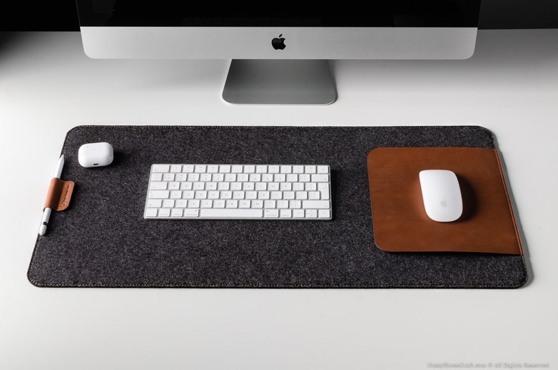 May include: A gray felt desk mat with a brown leather mouse pad and a white wireless keyboard. A white wireless mouse is on the brown leather mouse pad. A white wireless charging pad is on the left side of the mat.