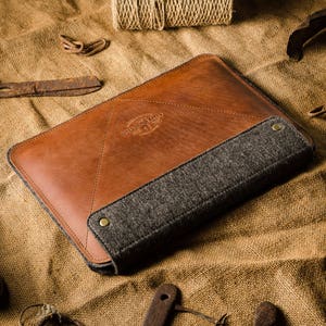 Puede incluir: Una funda para portátil de cuero marrón con forro de fieltro gris. La funda tiene un diseño cosido y un parche de cuero con el texto "Grey Horse Craft Co."