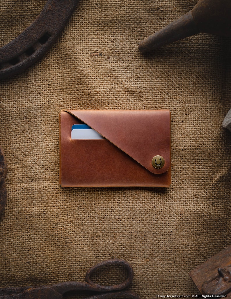 May include: A brown leather wallet with a flap closure and a horseshoe-shaped button. The wallet is on a burlap background.