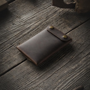 May include: A brown leather wallet with two brass snaps on the front. The wallet is lying on a wooden surface.