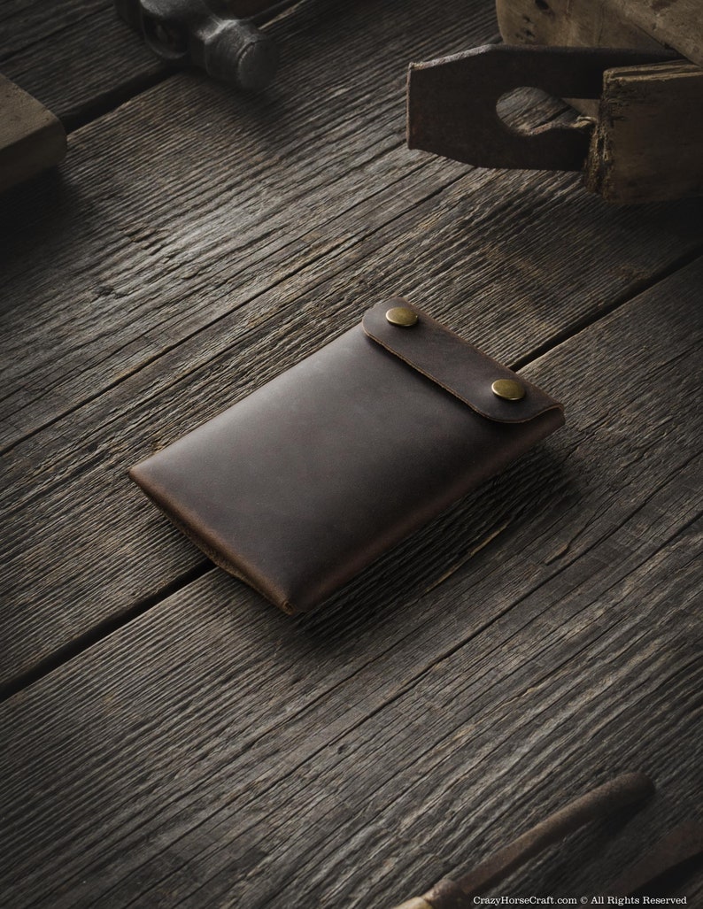 May include: A brown leather wallet with two brass snaps on the front. The wallet is lying on a wooden surface.
