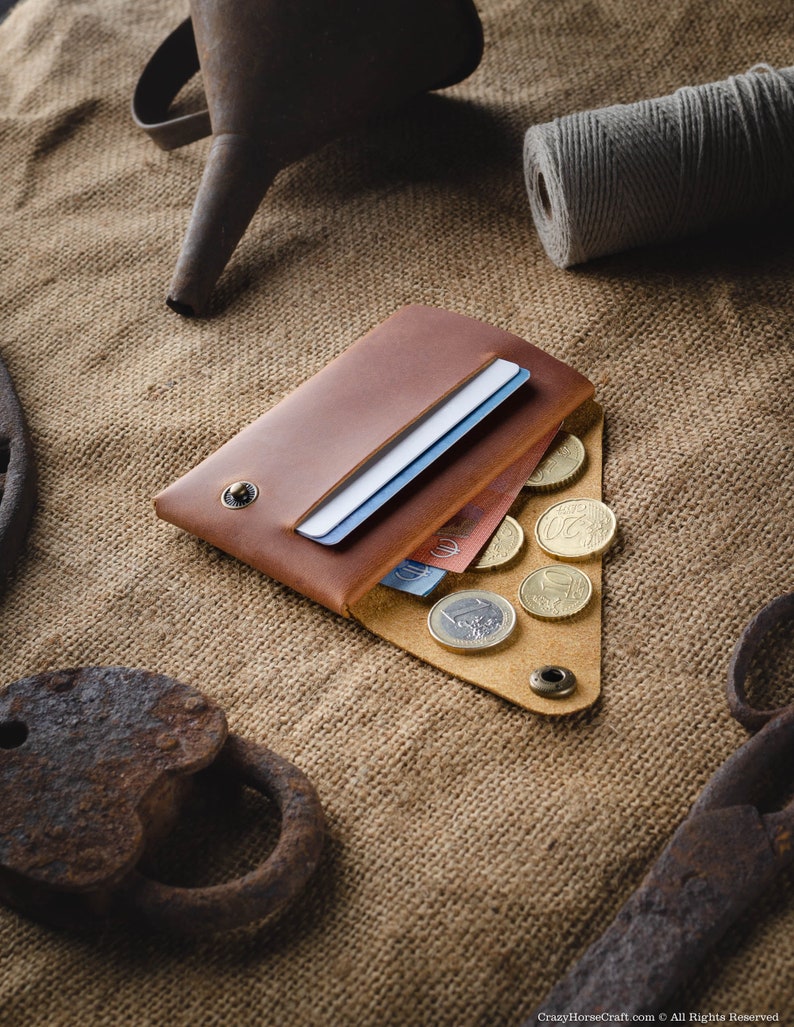 May include: A brown leather wallet with a flap open, revealing a few credit cards and euro coins. The wallet is lying on a burlap surface.
