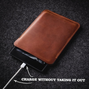 Puede incluir: Una funda de cuero marr&oacute;n para lector electr&oacute;nico con costuras visibles. El lector electr&oacute;nico es parcialmente visible, conectado a un cable de carga blanco. El texto dice "CHARGE WITHOUT TAKING IT OUT".