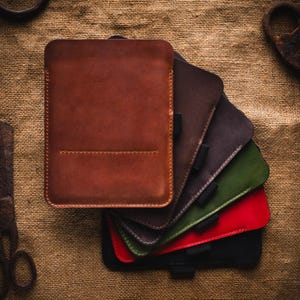 May include: A stack of leather tablet sleeves in various colors: brown, dark grey, green, red, and black. The top sleeve is a rich brown, with a stitched border. The sleeves are arranged on a textured, tan fabric.