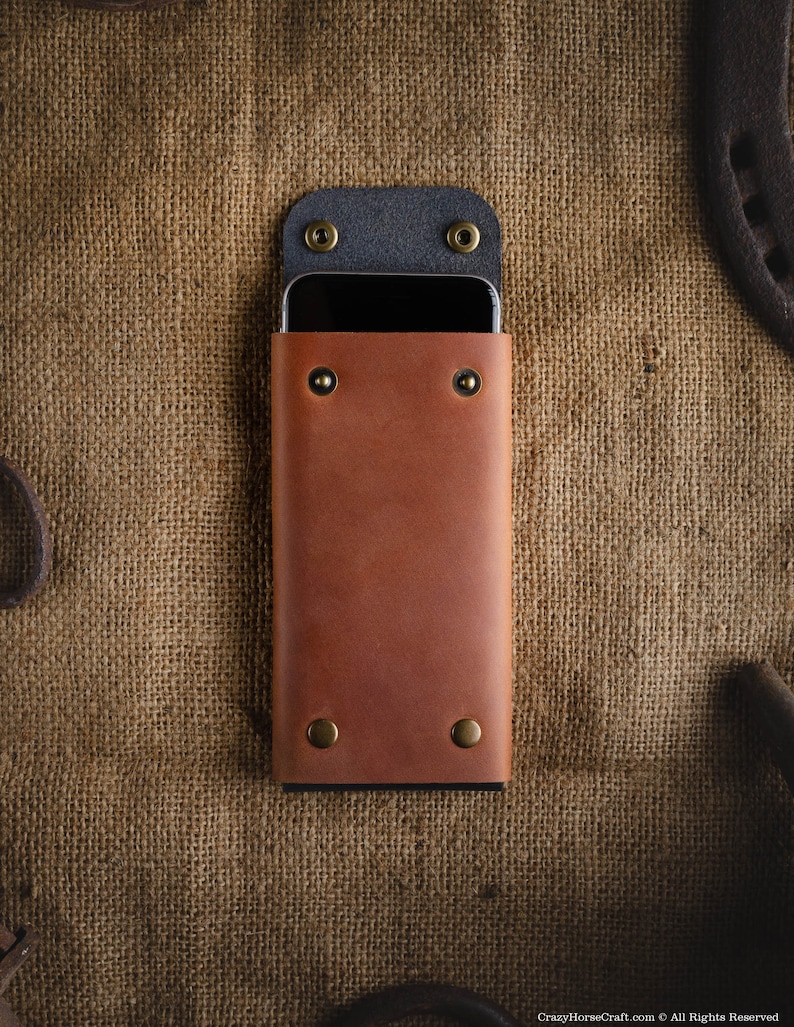May include: A brown leather phone case with a black flap and brass accents. The case is lying on a burlap background.