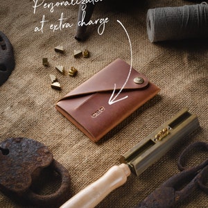 May include: A brown leather wallet with a brass button closure. The wallet is on a burlap background with a brass tool and metal letters. The text "Personalization available at extra charge" is written on the image.