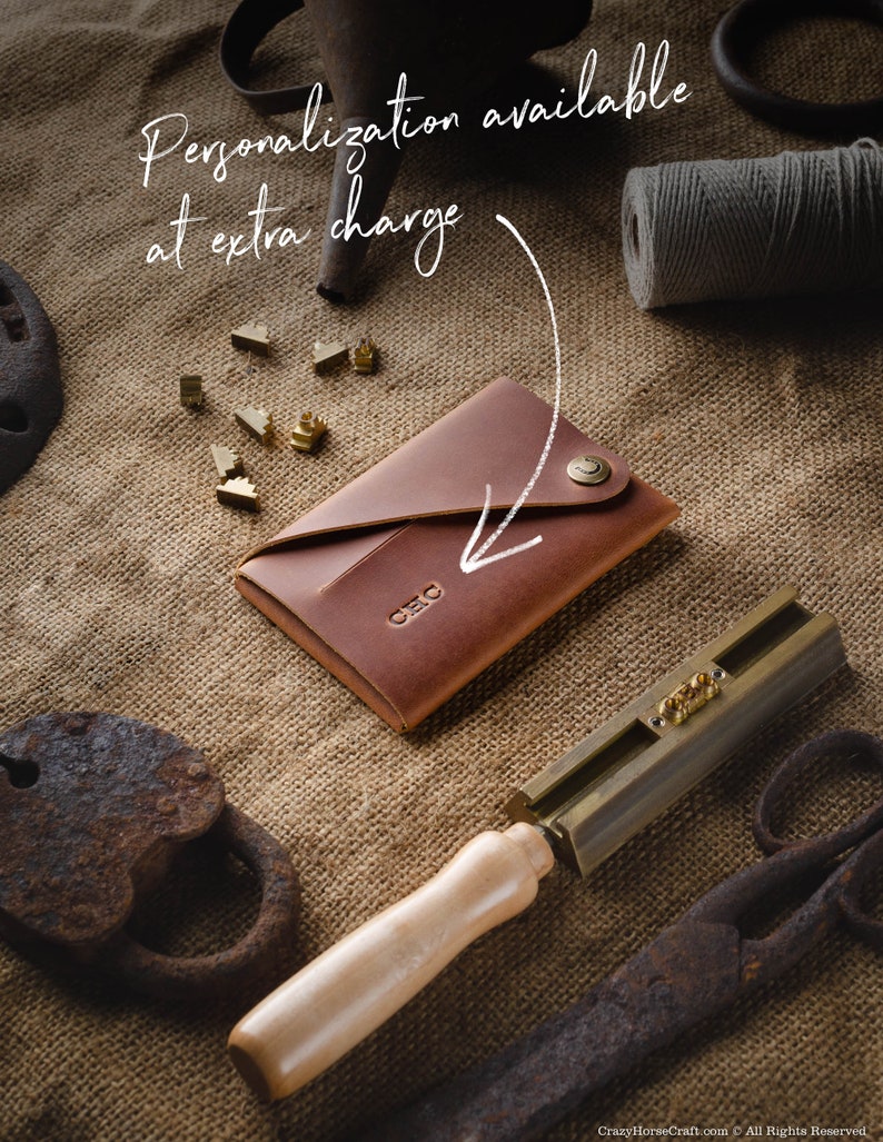 May include: A brown leather wallet with a brass button closure. The wallet is on a burlap background with a brass tool and metal letters. The text "Personalization available at extra charge" is written on the image.