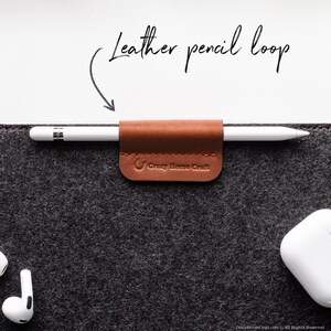 May include: A brown leather pencil loop with the text "Crazy Horse Craft" attached to a gray felt surface. A white Apple Pencil is inserted into the loop.