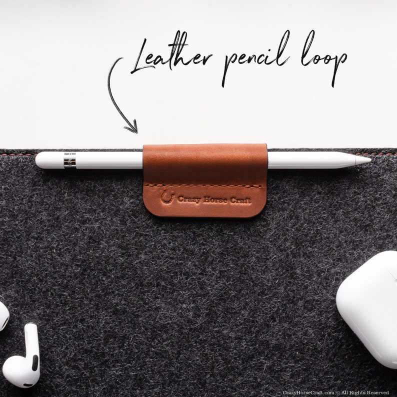 May include: A brown leather pencil loop with the text "Crazy Horse Craft" attached to a gray felt surface. A white Apple Pencil is inserted into the loop.