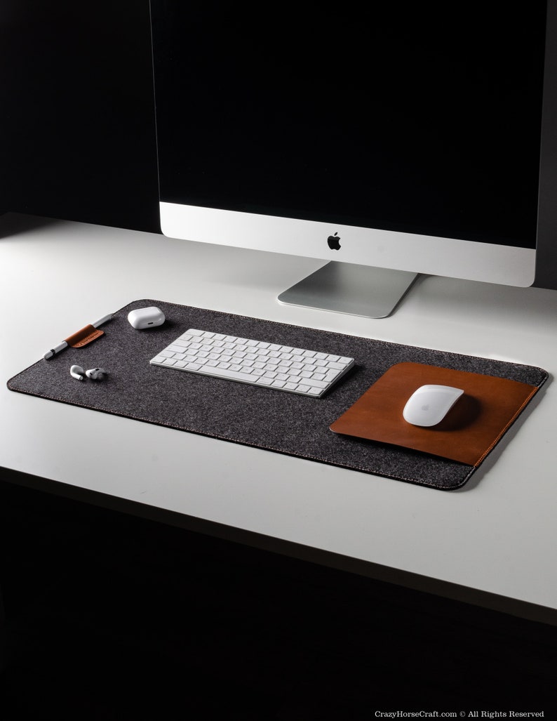 May include: A gray felt desk mat with a brown leather mouse pad and a white wireless keyboard and mouse. The mat is on a white desk.