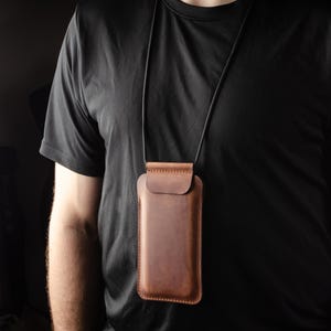 May include: A brown leather phone case with a black cord, worn around the neck of a person wearing a black t-shirt.