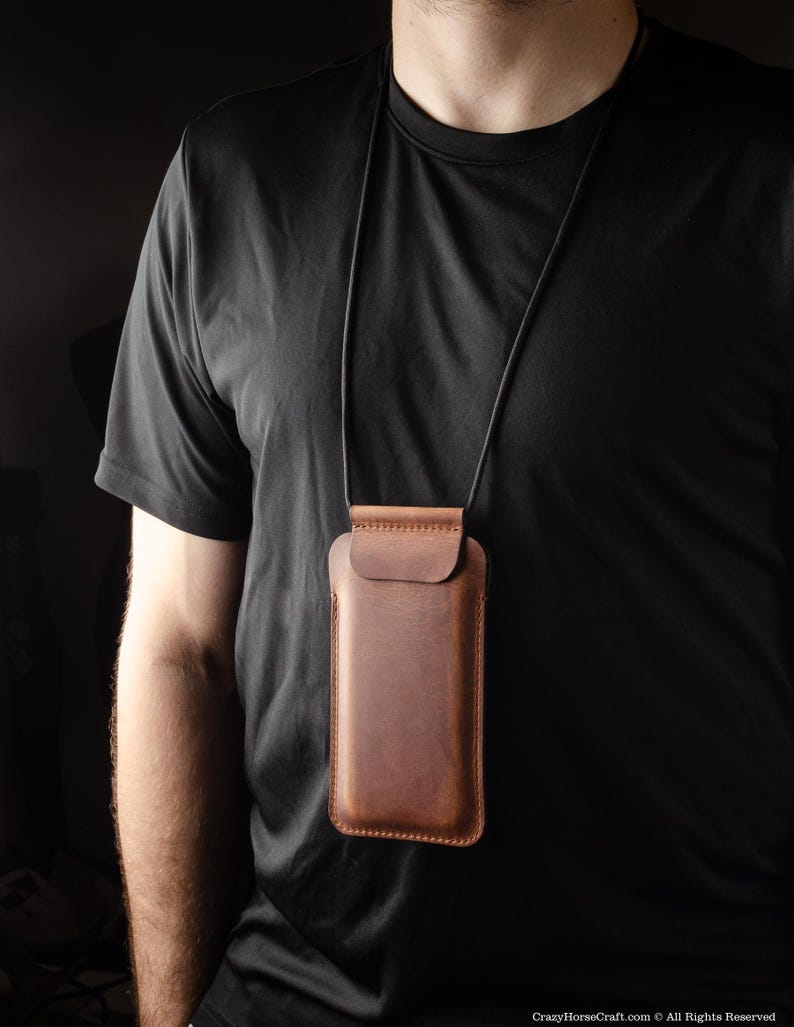 May include: A brown leather phone case with a black cord, worn around the neck of a person wearing a black t-shirt.