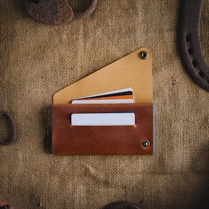 May include: A brown leather wallet with a flap and a snap closure. The wallet has a beige interior and is holding several credit cards. The wallet is lying on a burlap surface.