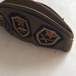 Puede incluir: Un sombrero de estilo militar marrón con tres parches bordados. Los parches presentan una estrella roja, espadas cruzadas y un insecto estilizado.