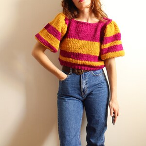 May include: A striped knit sweater in pink and yellow. The sweater has a round neckline and puffed sleeves.