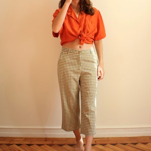 Pu&ograve; includere: Una camicia arancione a maniche corte annodata in vita, abbinata a pantaloni cropped a quadri verde oliva e bianchi. L'outfit &egrave; casual ed elegante, adatto per l'uso quotidiano.