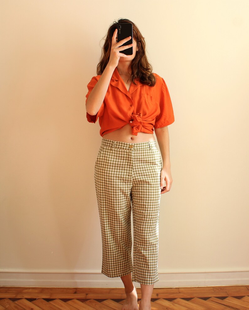 Pu&ograve; includere: Una camicia arancione a maniche corte annodata in vita, abbinata a pantaloni cropped a quadri verde oliva e bianchi. L'outfit &egrave; casual ed elegante, adatto per l'uso quotidiano.