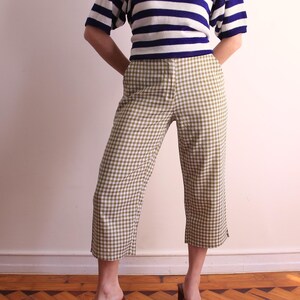 Pu&ograve; includere: Una persona indossa una maglietta a maniche corte a righe blu e bianche e pantaloni cropped a quadri verde oliva e bianchi. I pantaloni hanno una gamba larga e sono a met&agrave; polpaccio. La persona &egrave; in piedi su un pavimento di legno.