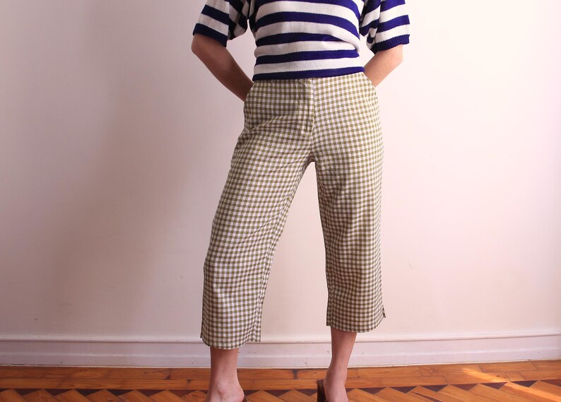 Pu&ograve; includere: Una persona indossa una maglietta a maniche corte a righe blu e bianche e pantaloni cropped a quadri verde oliva e bianchi. I pantaloni hanno una gamba larga e sono a met&agrave; polpaccio. La persona &egrave; in piedi su un pavimento di legno.
