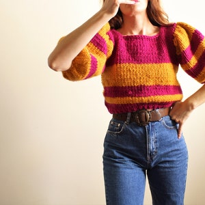 May include: A striped sweater with a pink and yellow colour scheme. The sweater has a square neckline and puffed sleeves. The sweater is cropped and is being worn with a brown belt and blue jeans.
