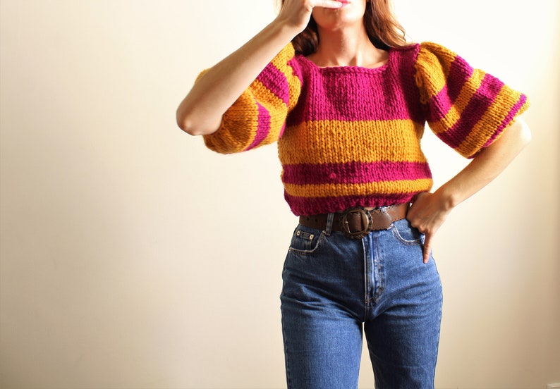 May include: A striped sweater with a pink and yellow colour scheme. The sweater has a square neckline and puffed sleeves. The sweater is cropped and is being worn with a brown belt and blue jeans.