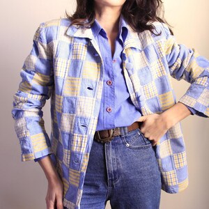 Pode incluir: Uma jaqueta azul e amarela xadrez com uma camisa azul de botões por baixo. A jaqueta tem um design de patchwork e é usada sobre uma calça jeans azul.