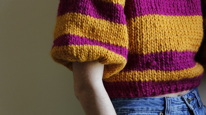May include: A striped jumper with yellow and pink horizontal stripes. The jumper has a cropped length and a relaxed fit.