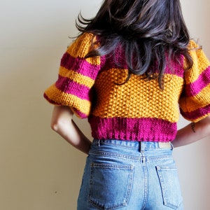 May include: A cropped sweater with yellow and pink horizontal stripes and puffed sleeves. The sweater is made of a soft, textured yarn.