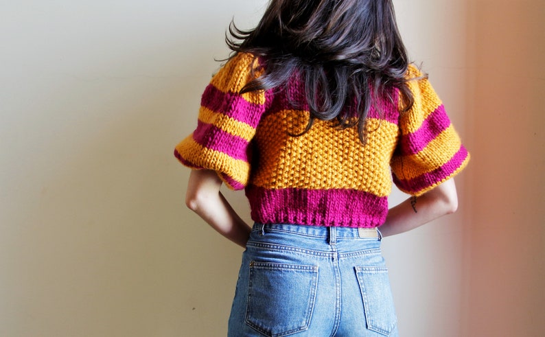 May include: A cropped sweater with yellow and pink horizontal stripes and puffed sleeves. The sweater is made of a soft, textured yarn.