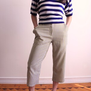 Pu&ograve; includere: Una persona indossa una maglietta a maniche corte a righe blu e bianche e pantaloni cropped a quadri verde oliva e bianchi. I pantaloni hanno tasche e sono al ginocchio. L'outfit &egrave; casual ed elegante.