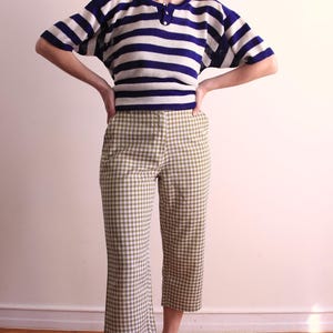 Pu&ograve; includere: Un completo a due pezzi con un top a maniche corte a righe blu navy e bianche con scollo a bottoni, abbinato a pantaloni a quadri verde oliva e bianchi. L'outfit &egrave; in stile vintage.