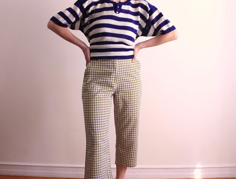 Pu&ograve; includere: Un completo a due pezzi con un top a maniche corte a righe blu navy e bianche con scollo a bottoni, abbinato a pantaloni a quadri verde oliva e bianchi. L'outfit &egrave; in stile vintage.