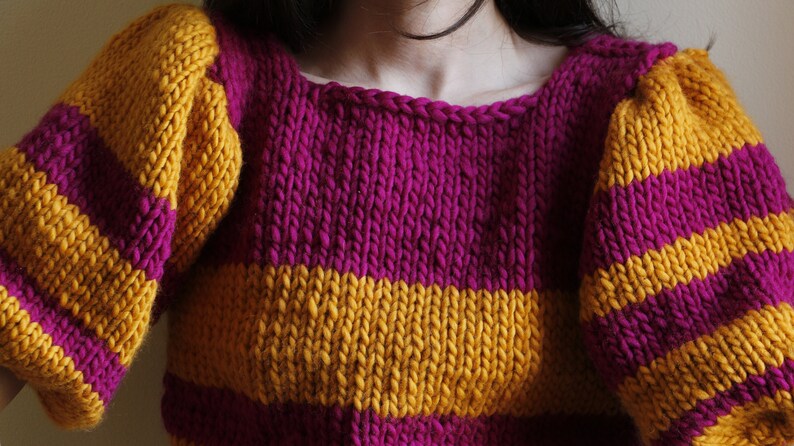 May include: A striped knitted jumper with yellow and purple stripes. The jumper has a round neckline and long sleeves.