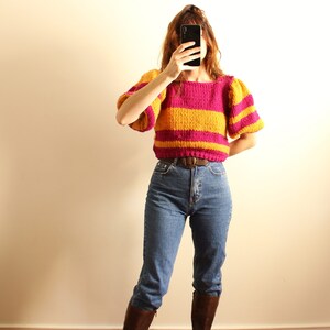 May include: A woman wearing a pink and yellow striped jumper with puffed sleeves, blue jeans, and brown boots. She is holding a black smartphone in front of her face.