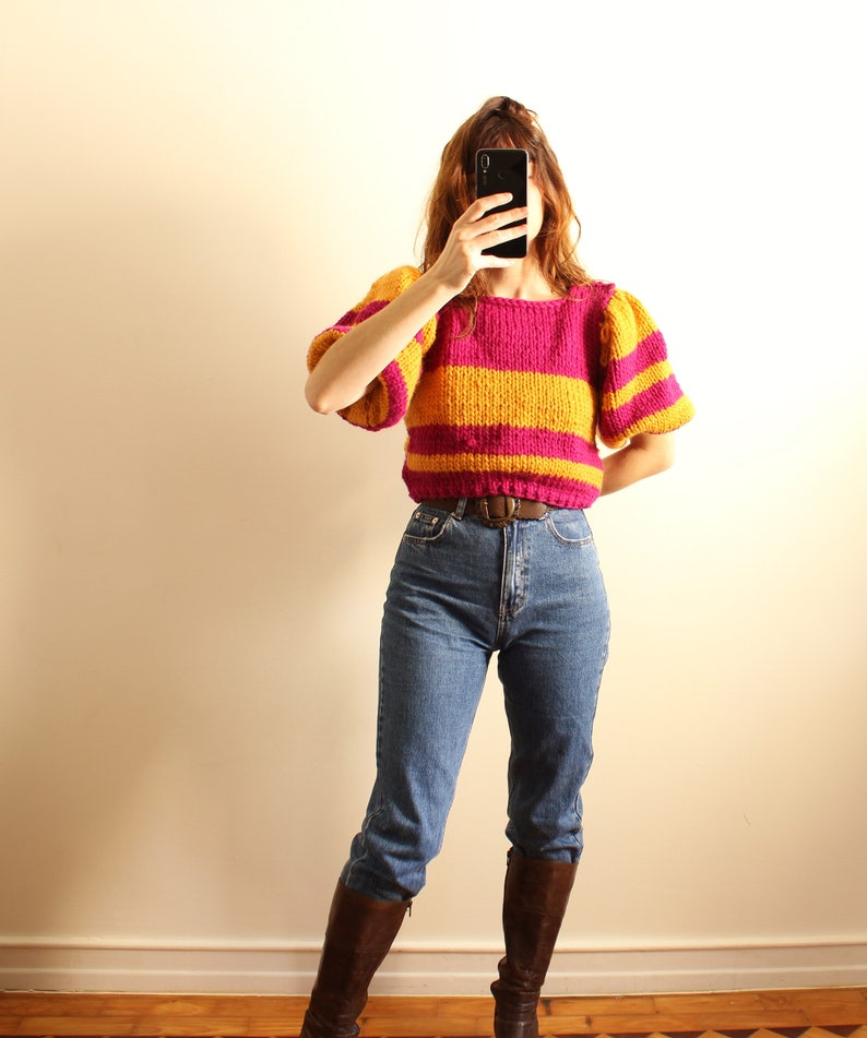 May include: A woman wearing a pink and yellow striped jumper with puffed sleeves, blue jeans, and brown boots. She is holding a black smartphone in front of her face.