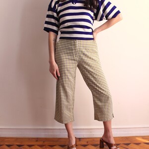 Pu&ograve; includere: Una persona indossa una camicia a maniche corte a righe blu navy e bianche e pantaloni cropped a quadri verde oliva e bianchi. La persona indossa anche sandali con tacco marroni. L'outfit &egrave; in stile vintage.