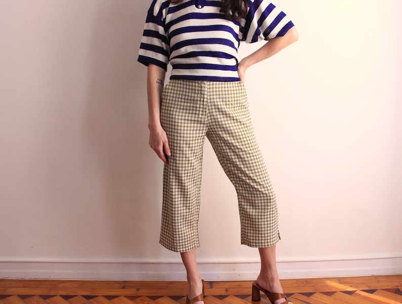Pu&ograve; includere: Una persona indossa una camicia a maniche corte a righe blu navy e bianche e pantaloni cropped a quadri verde oliva e bianchi. La persona indossa anche sandali con tacco marroni. L'outfit &egrave; in stile vintage.