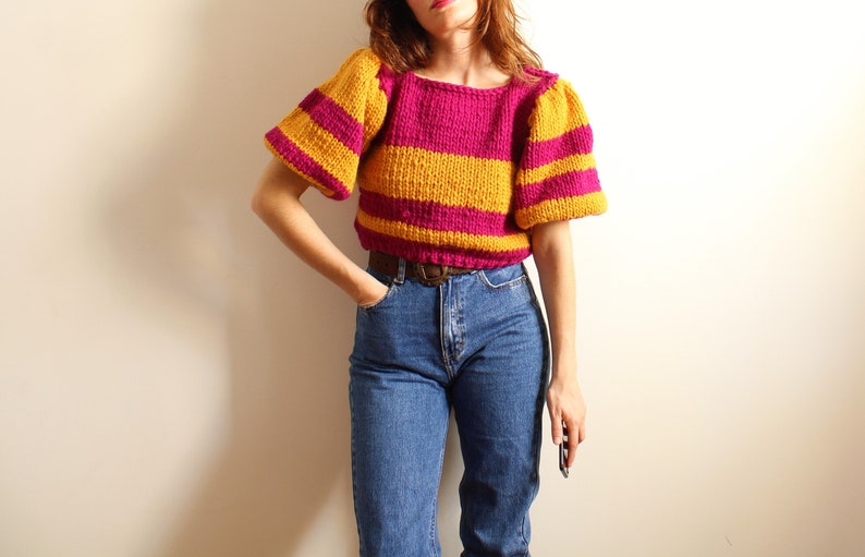 May include: A striped knit sweater in pink and yellow. The sweater has a round neckline and puffed sleeves.